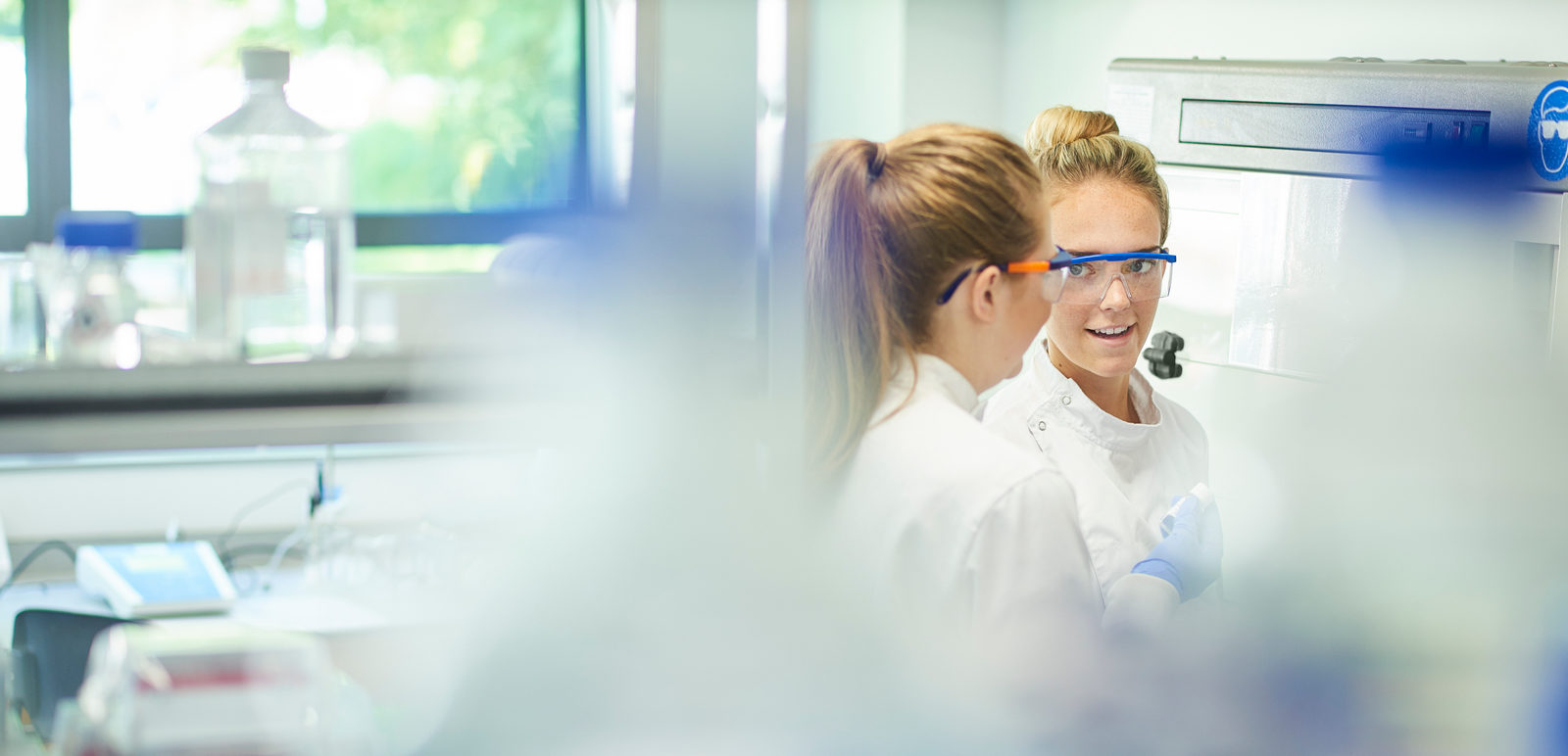 To kvinnelige laboratoriearbeidere. I forgrunnen noen defokuserte hyller og laboratorieglass. Fotografi.