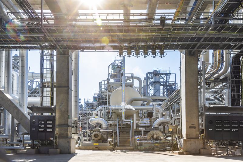 Et industrielt anlegg, sannsynligvis et raffineri eller kjemisk fabrikk, med ulike rør, tanker og maskineri. Det er flere nivåer med metallstrukturer og gangveier, sammen med elektriske paneler på sidene. Solen skinner sterkt ovenfra, og skaper lysrefleksjoner i bildet. Foto.