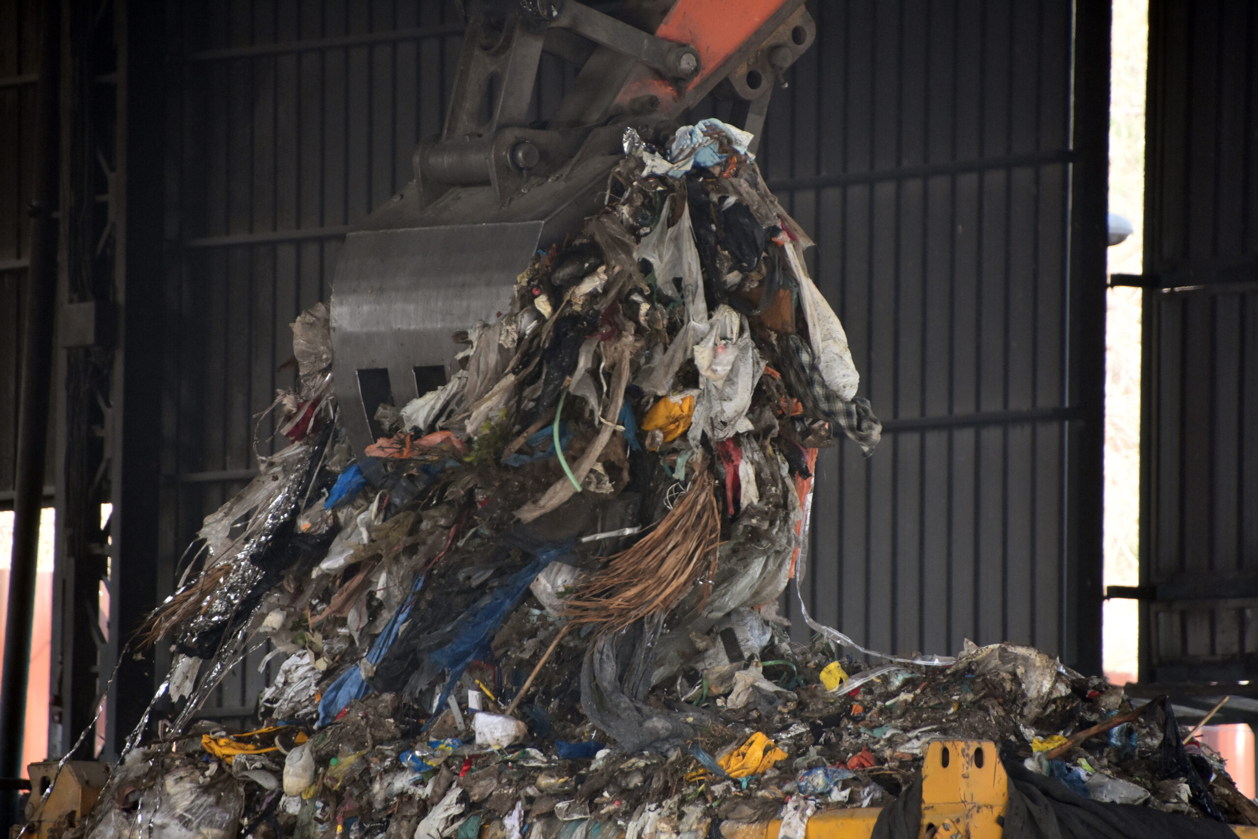 En stor industriell klo løfter en haug med blandede avfallsmaterialer, inkludert plast, metall og annet avfall, inne i det som ser ut til å være et avfallshåndterings- eller gjenvinningsanlegg. Foto.