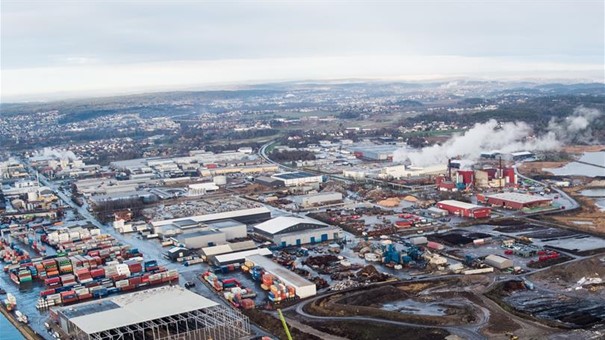 Luftfoto av et industriområde med mange lagerbygninger, fabrikker og lagringsanlegg. Flere stablede fraktcontainere og områder med synlig røyk eller damp. I bakgrunnen ses boligområder og et landskap som strekker seg mot horisonten. Foto.
