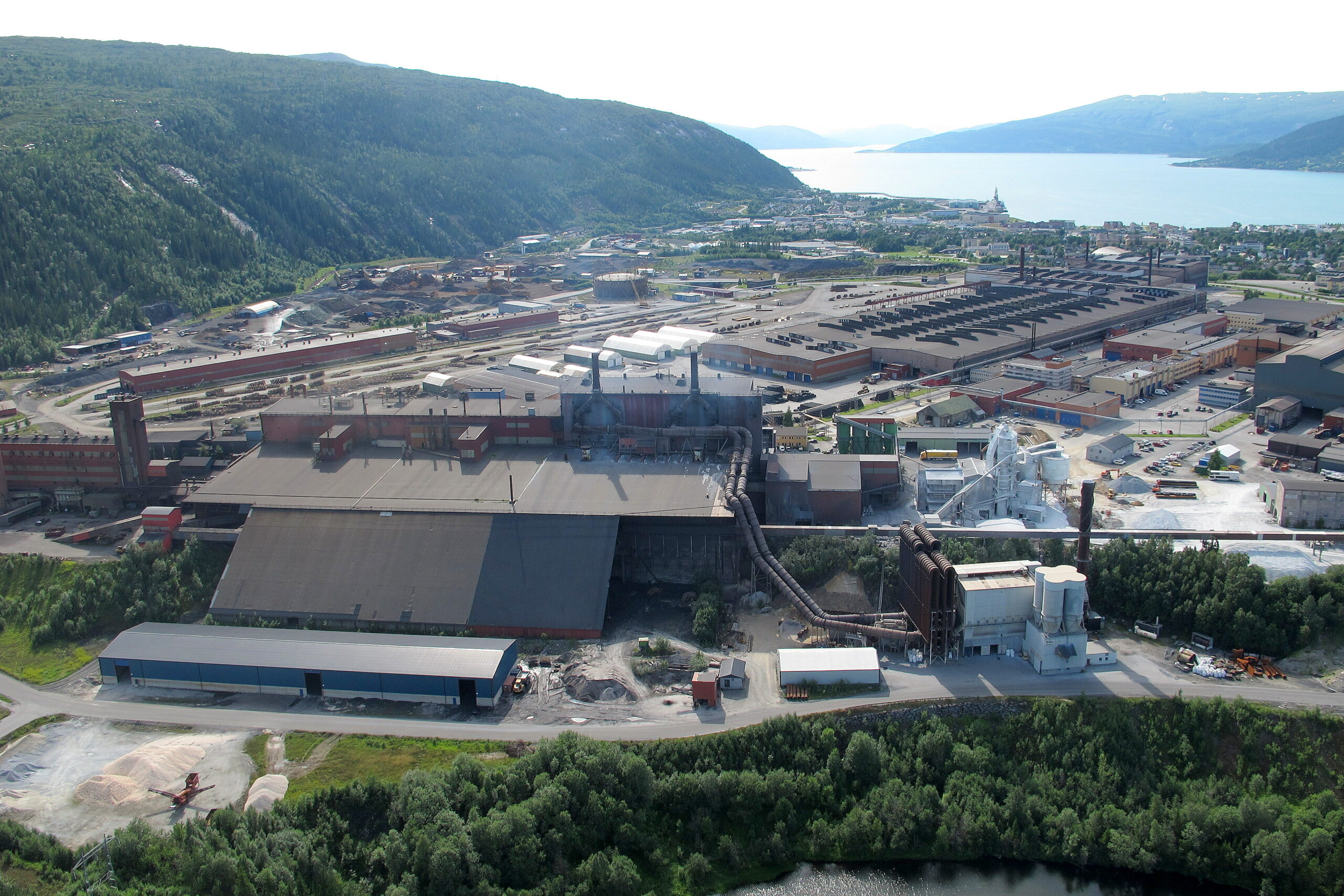 Flyfoto av et industriområde omgitt av frodig grønt landskap og åser. Komplekset inkluderer store bygninger, lagerhus og ulike industrielle strukturer. Det er også veier og mindre bygninger innenfor komplekset. I bakgrunnen er det fjord eller hav med mer land synlig bortenfor. Foto.