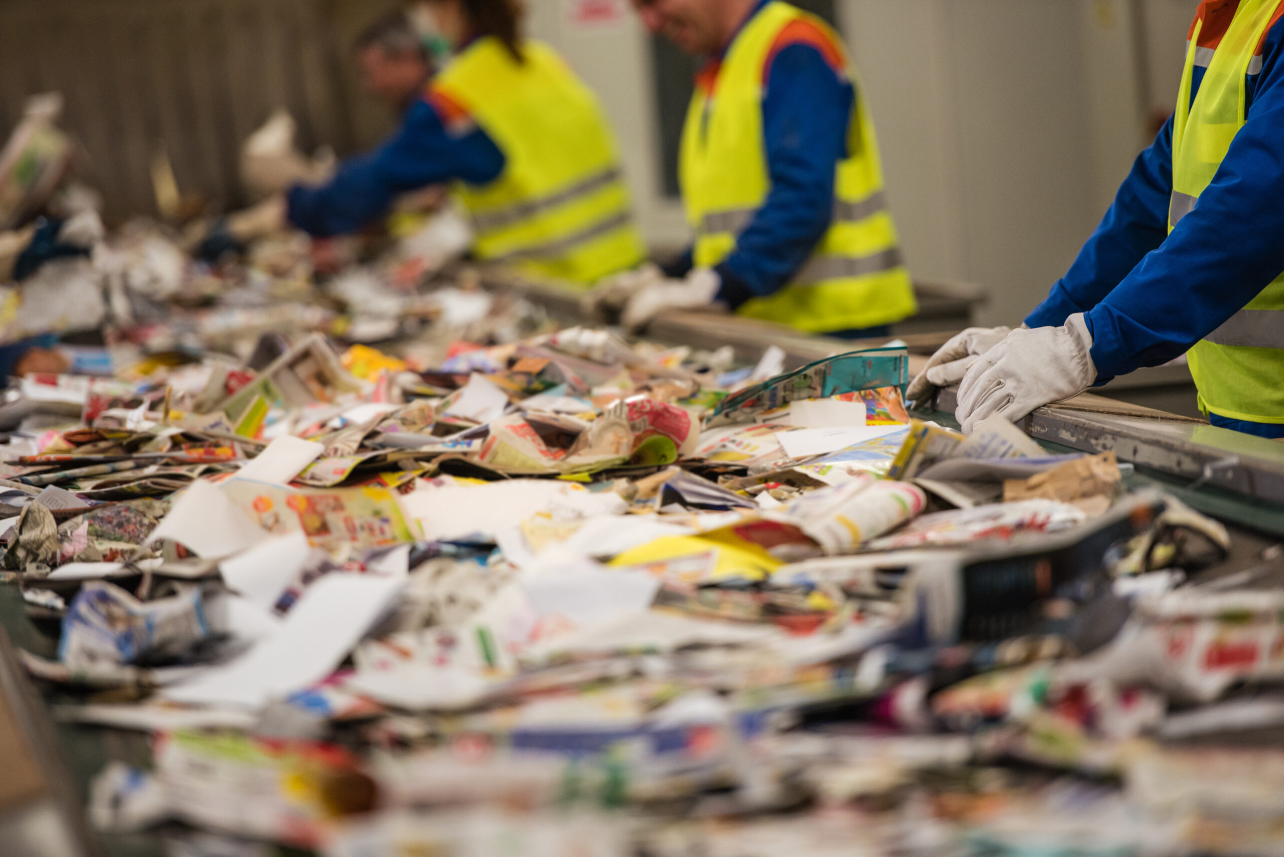 Flere arbeidere iført synlighetsvester og hansker sorterer gjennom en stor mengde blandet papir og pappmaterialer på et transportbånd. Foto.