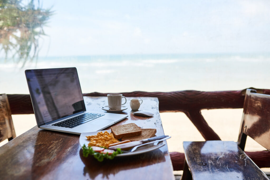 Et trebord og benk utendørs med en bærbar datamaskin, en kopp kaffe og en tallerken med mat som inkluderer toast og eggerøre. Bordet er plassert med utsikt over havet i bakgrunnen. Foto.