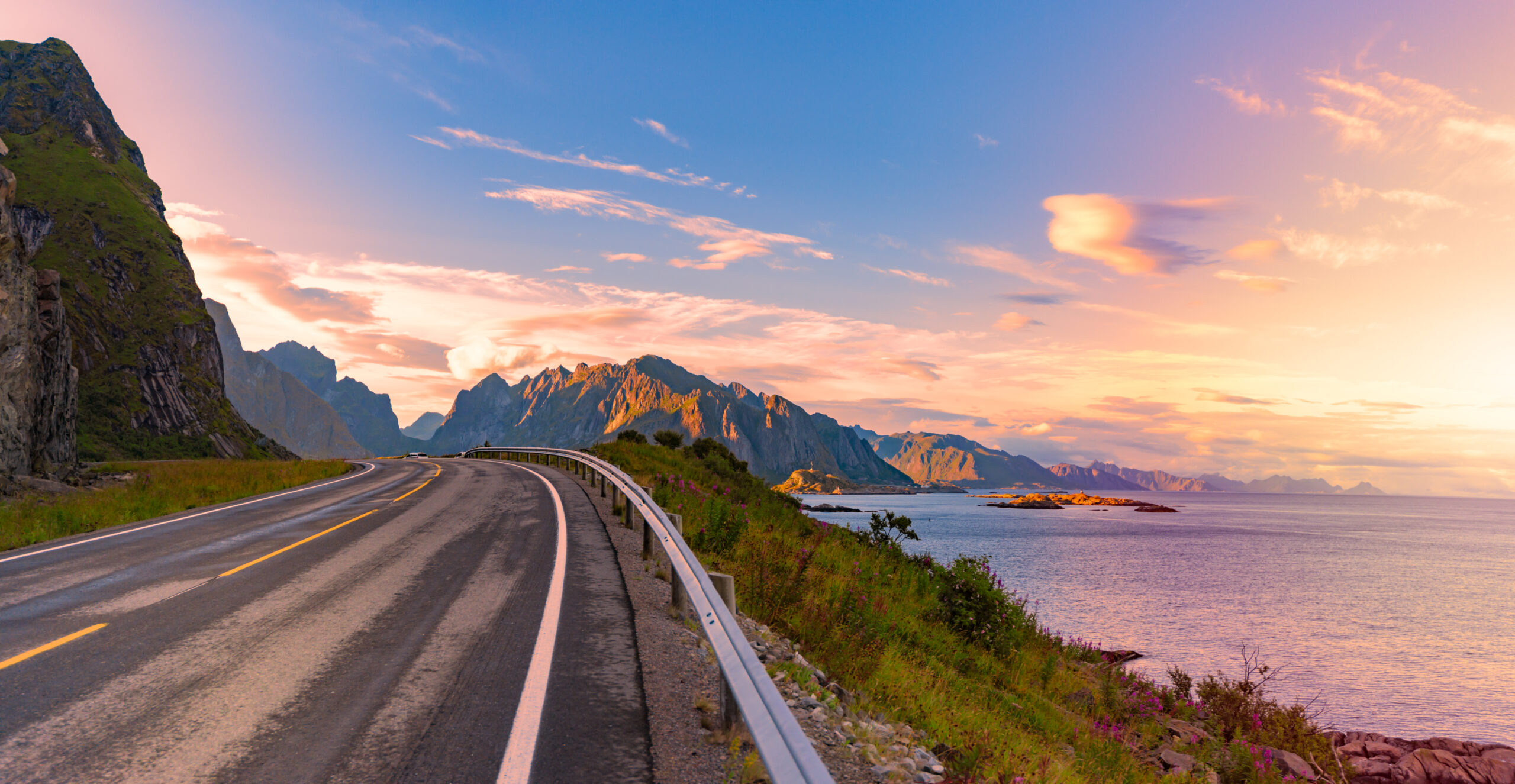 En naturskjønn, tom, kystvei i Lofoten som svinger langs kanten av en havet, med fjell i bakgrunnen. Himmelen har nyanser av rosa, oransje og blått, noe som indikerer enten soloppgang eller solnedgang. Veien har et rekkverk på høyre side og er omgitt av grøntområder og steinete terreng. Foto.