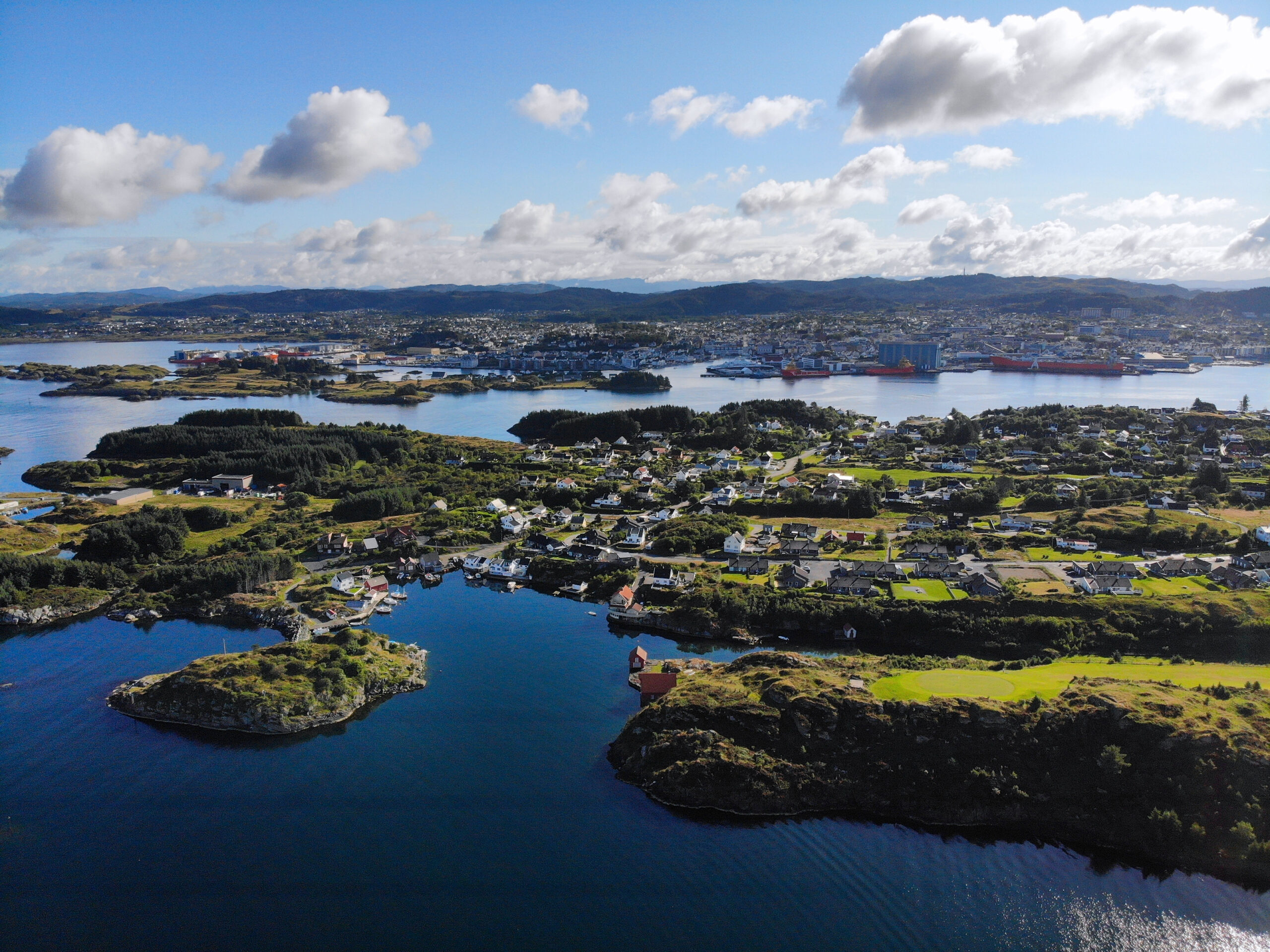 Et luftfoto av et kystlandskap med mange små øyer og halvøyer omgitt av vann nær Haugesund by. Området er prikket med hus, grønne marker og skogsområder. I bakgrunnen er det et større landområde med tettere bebyggelse og flere store skip som ligger til kai langs kysten. Himmelen er delvis skyet med spredte hvite skyer. Foto.
