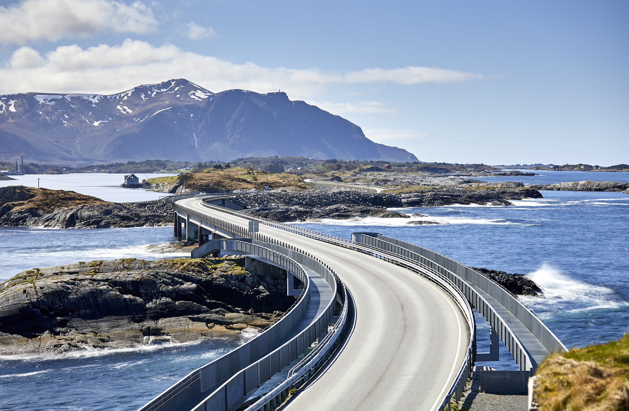 Norsk landskap med holmer, skjær, hav, bølger og fjell i det fjerne. Midt i bildet er den berømte Atlanterhavsbroen. Foto.